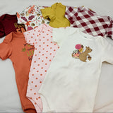 A flat lay of six baby bodysuits in various patterns and colors, including solid white, pink polka dots, orange, red and white gingham, yellow, and a floral print, arranged on a white fabric background.